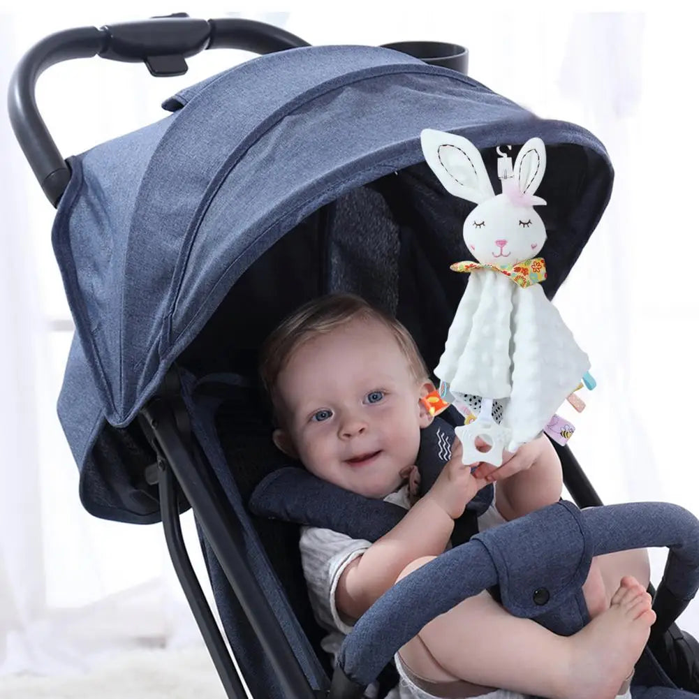 A baby in a stroller holding a plush bunny comforter with colorful tags, wearing a white outfit.