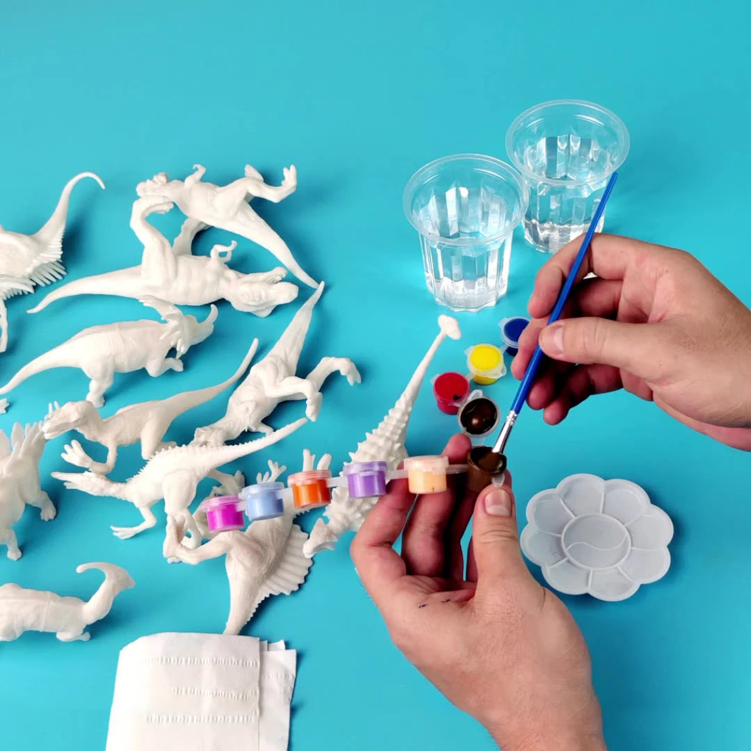 Unpainted dinosaur models in white, paint pots in various colors, and brushes on a blue background.