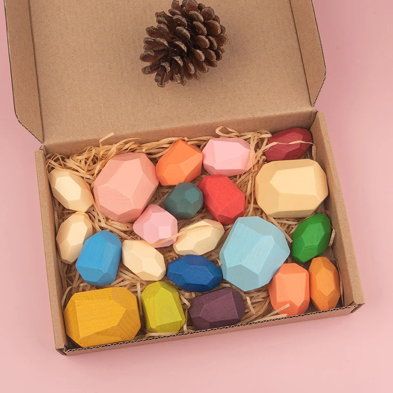 Colorful wooden sorting stones in a cardboard box with straw, including various shapes and sizes, and a pine cone on top.