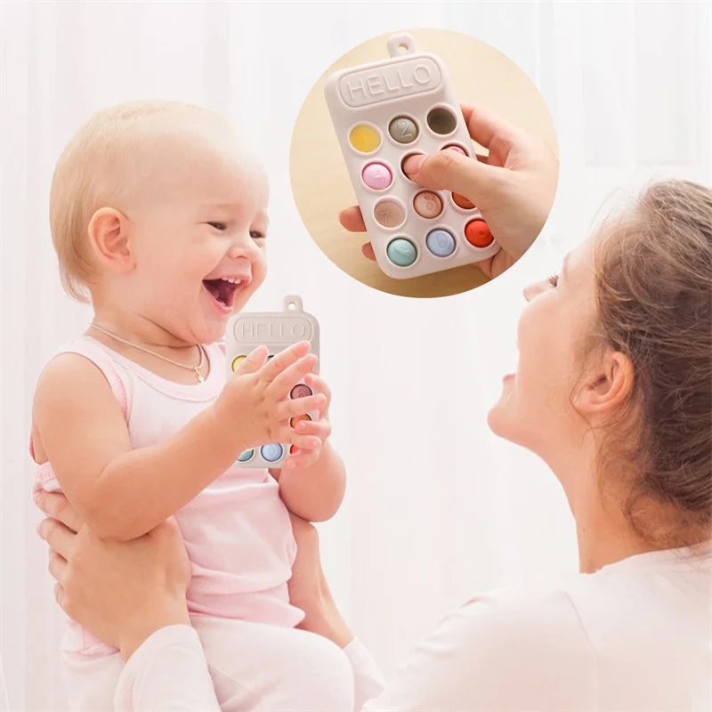 A baby holding a silicone cell phone teether toy with colorful buttons, smiling at a woman holding them.