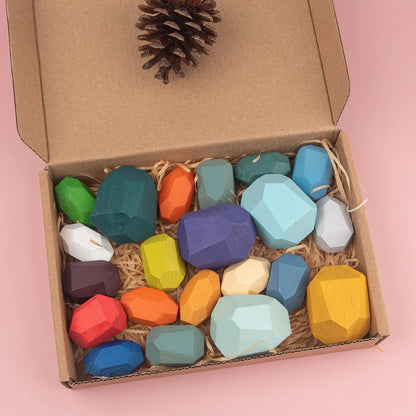 Colorful wooden sorting stones in a cardboard box with straw, includes various shapes and a pinecone on top.