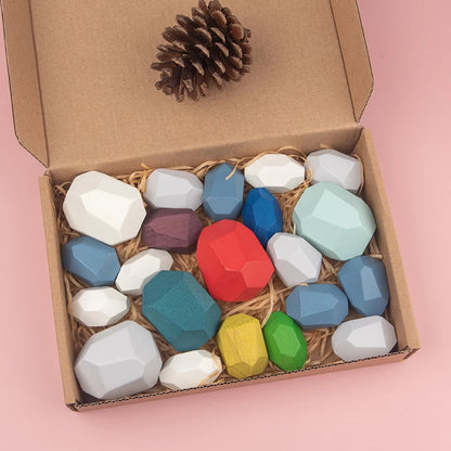 Colorful wooden sorting stones in a cardboard box with straw, includes various shapes and a pinecone on top.