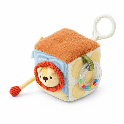 A baby sensory cube toy with a lion face, orange plush top, blue striped side, and a clear ring rattle, shown with dimension labels and a small inset instruction graphic on a white background.