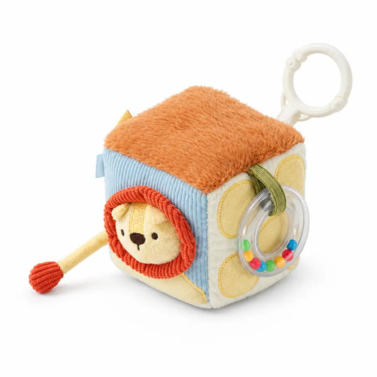 A baby sensory cube toy with a lion face, orange plush top, blue striped side, and a clear ring rattle, shown with dimension labels and a small inset instruction graphic on a white background.