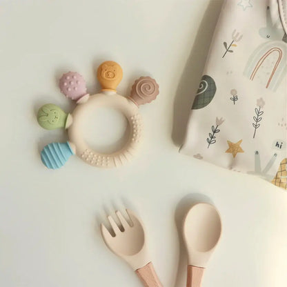 Silicone baby teether with colorful beads, surrounded by baby utensils on a patterned cloth.