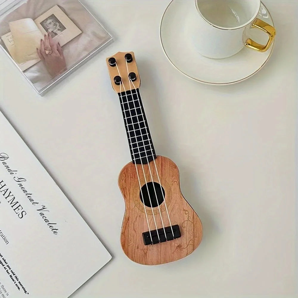Mini classical ukulele guitar toy in light wood finish, placed on a flat surface with a cup and books.