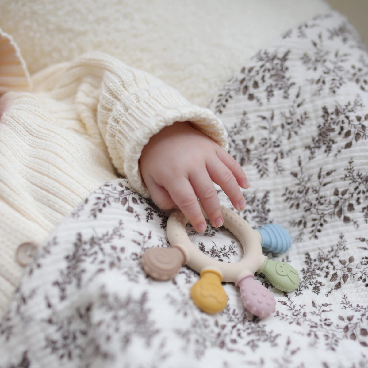 Silicone baby teether with colorful chew beads, held by a baby in a cozy sweater on a patterned blanket.