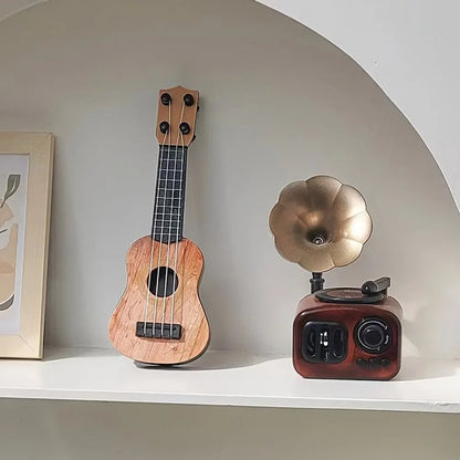 Mini classical ukulele guitar toy in wood finish, displayed on a shelf with a vintage gramophone and framed art.